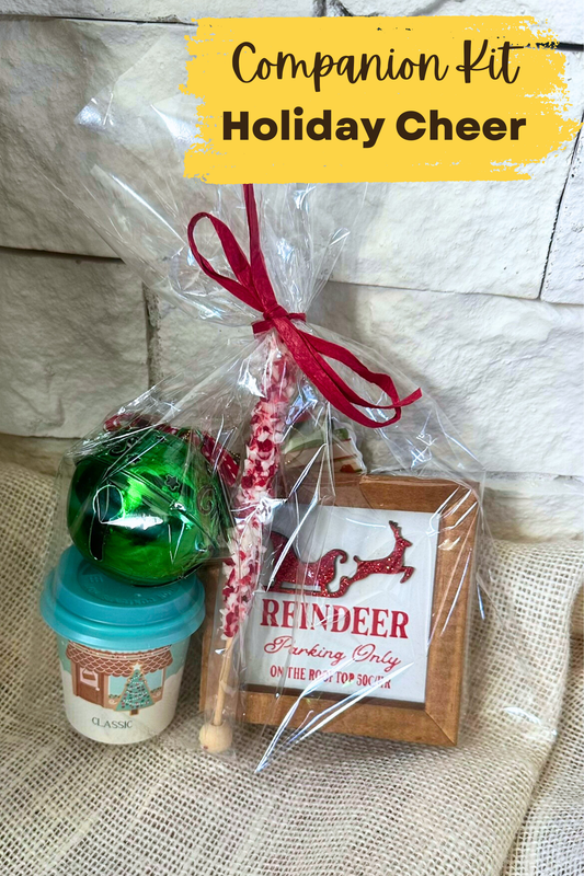 Cellophane bag with wooden holiday decoration, colorful bell ornament, hot cocoa in a small coffee cup, cocoa stir stick, and holiday bookmark