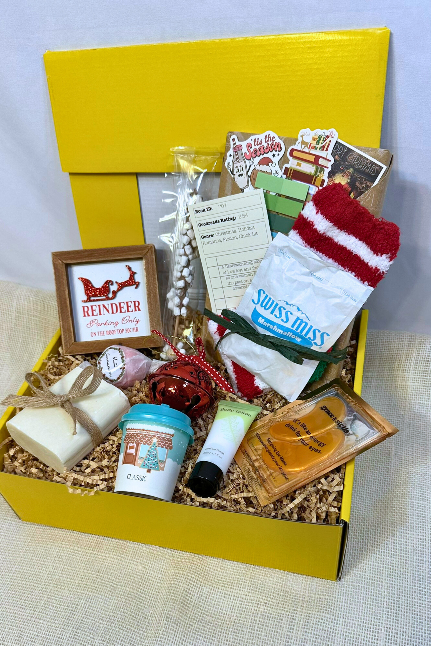Gift box with various holiday items including handmade bar soap, body lotion, holiday decorations, hot chocolate, and blind date with a book.