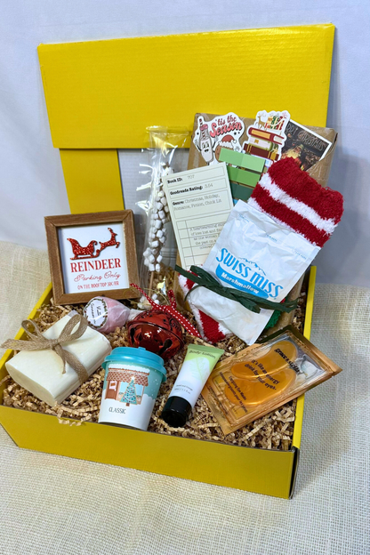 Gift box with various holiday items including handmade bar soap, body lotion, holiday decorations, hot chocolate, and blind date with a book.