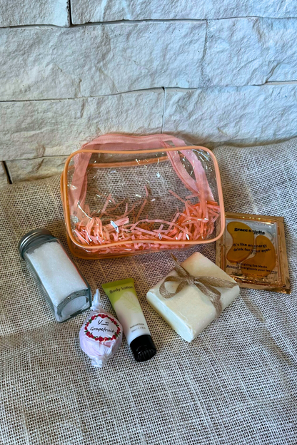 Clear zipper bag with pink trim with handmade bar soap, eye mask, body lotion, bath salts, and bath bomb.