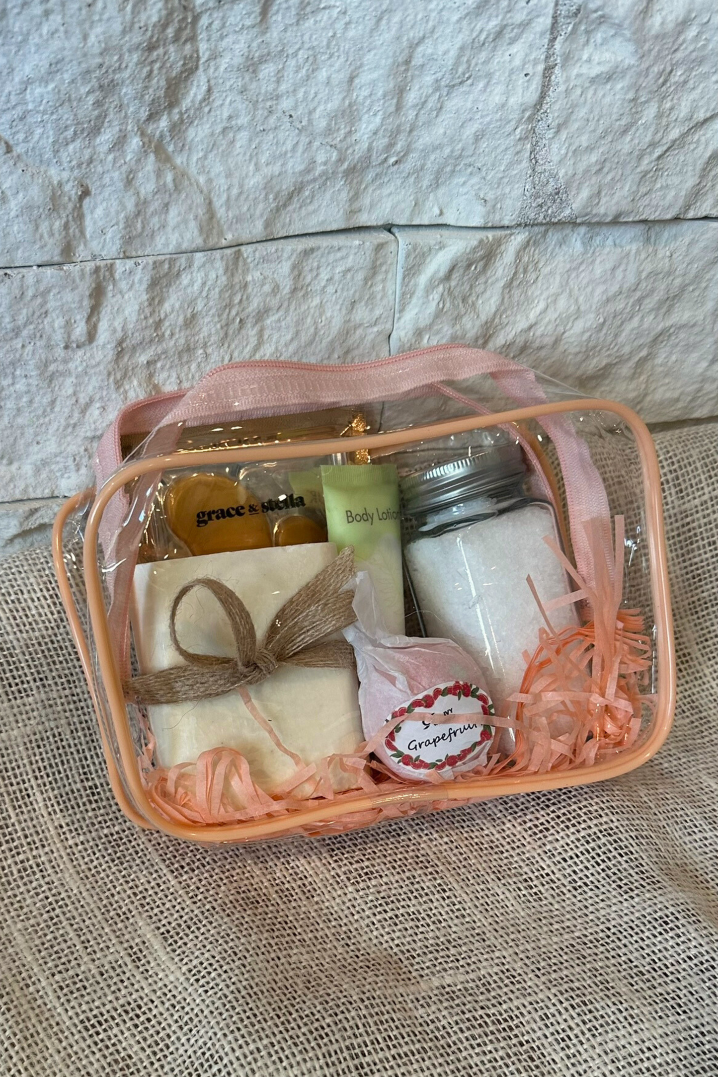 Clear zipper bag with pink trim with handmade bar soap, eye mask, body lotion, bath salts, and bath bomb inside